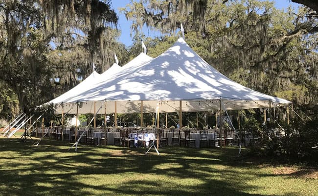 White event tent set up in a park with tables and chairs underneath, surrounded by trees draped with Spanish moss.