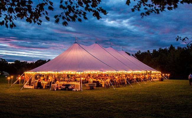 Large tent illuminated by string lights at dusk in an open grassy field, set up for an event with people visible under it.