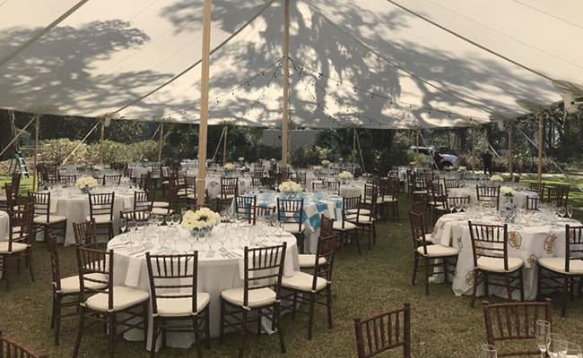 Outdoor event setup with multiple round dining tables under a large white tent, surrounded by a grassy area.