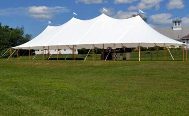 Large white event tent set up on a grassy field under a clear sky, with a small building partially visible in the background.