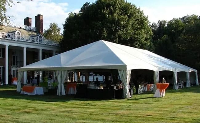 Large white event tent set up on a grassy lawn with tables and chairs visible inside, in front of an elegant house with balconies.