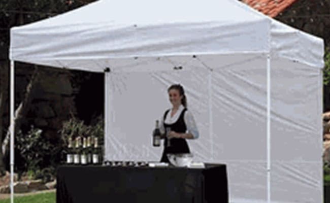 A waitress stands behind a table under a white canopy, preparing beverages with bottles and glasses in front of her.