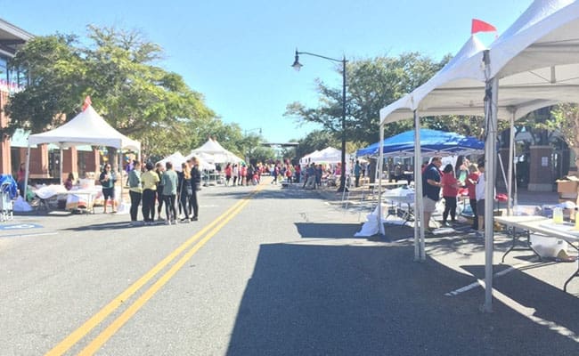 Street fair scene with people browsing vendor booths along a sunny, tree-lined street.