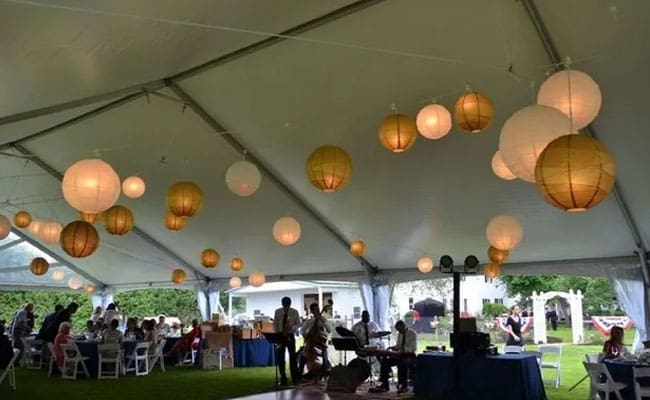 Event under a large tent with hanging paper lanterns, featuring seated guests, tables, and a DJ booth, set in a grassy area.