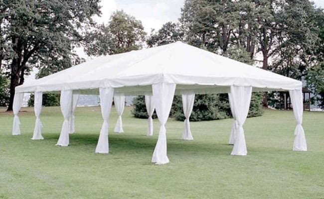 A white marquee tent with open sides and draped curtains, set up on a grassy area surrounded by trees.