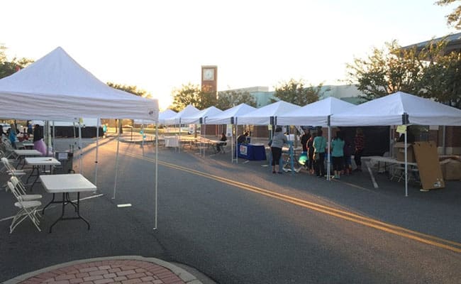 White tents set up along a street for an outdoor event, with a few people browsing and a clock tower in the background.