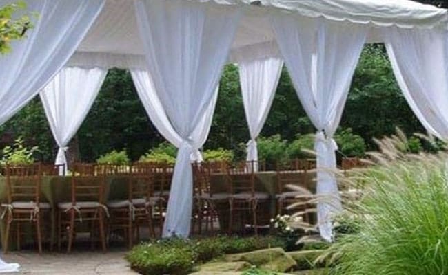 Outdoor wedding setup with white drapes and wooden chairs arranged under a canopy, surrounded by lush greenery.