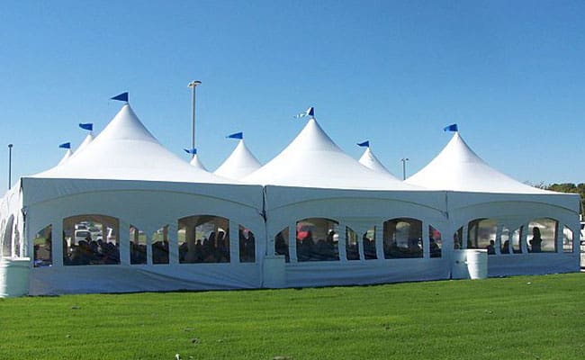 White event tents with blue flags and open sides set up on a grassy field under clear blue sky.