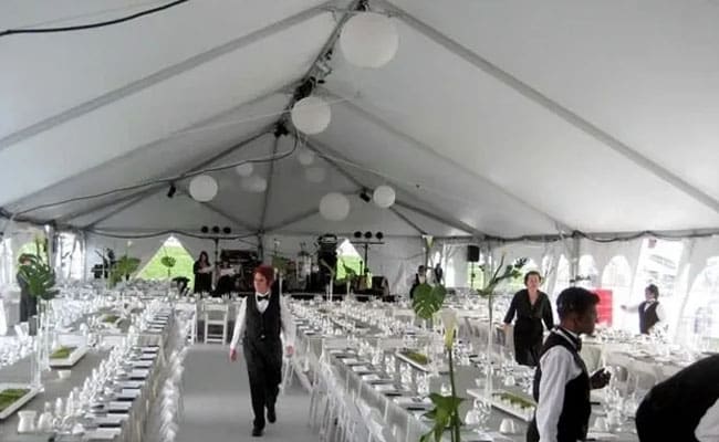 Large white event tent with rows of set dining tables, white chairs, and hanging paper lanterns, staff arranging settings.