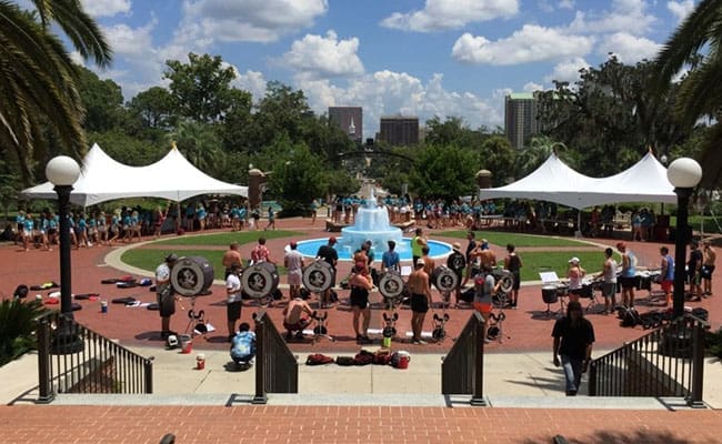 Outdoor fitness event at a park with people exercising near a fountain, under sunny skies, flanked by white tents and lampposts.