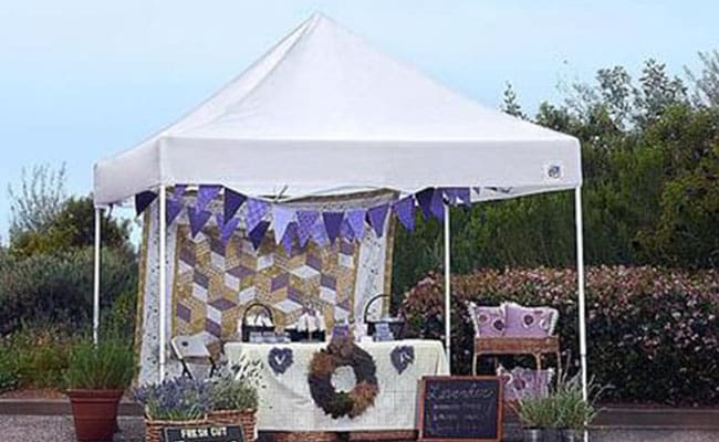 An outdoor market stall under a white canopy decorated with purple bunting, showcasing various home decor items and a "lavender" themed sign.