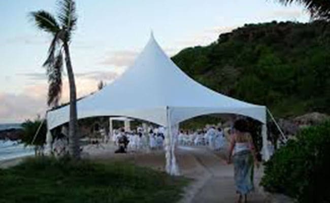 A white tent at a beachside wedding reception with guests in formal white attire, palms nearby, during twilight.