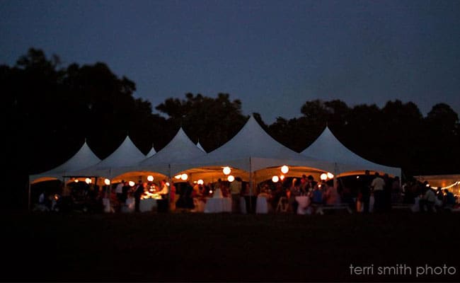 Nighttime view of an outdoor event under lit tents, with people gathered and a dark, wooded background.