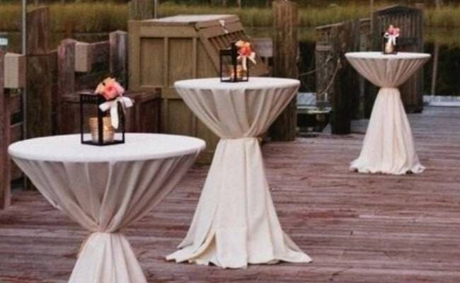 Cocktail tables with white drapes and small flower arrangements on a wooden deck, surrounded by rustic crates and barrels.