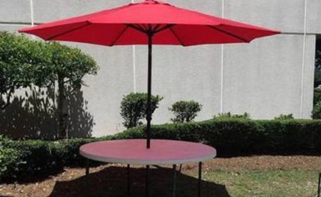 Red umbrella shading a round table, set against a building wall with small bushes under bright sunlight.