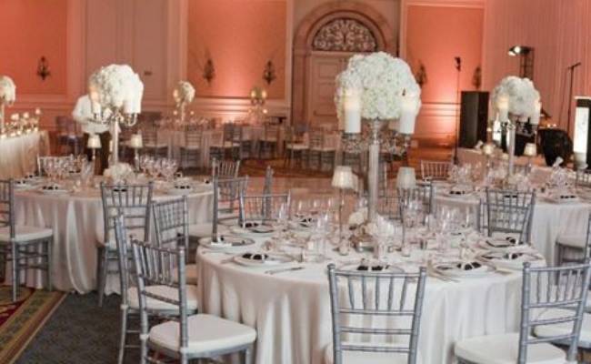 Elegant banquet hall set up for a formal event, featuring round tables with white linens and floral centerpieces, and silver chiavari chairs.