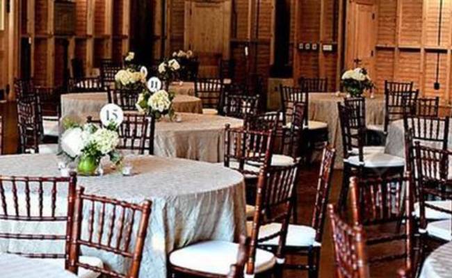 Elegant event setup featuring round tables with beige tablecloths, wooden chairs, and white floral centerpieces in a room with wooden walls and floors.