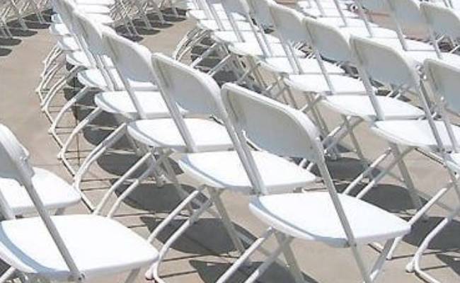 Rows of empty white folding chairs arranged outdoors, casting shadows on a concrete surface.