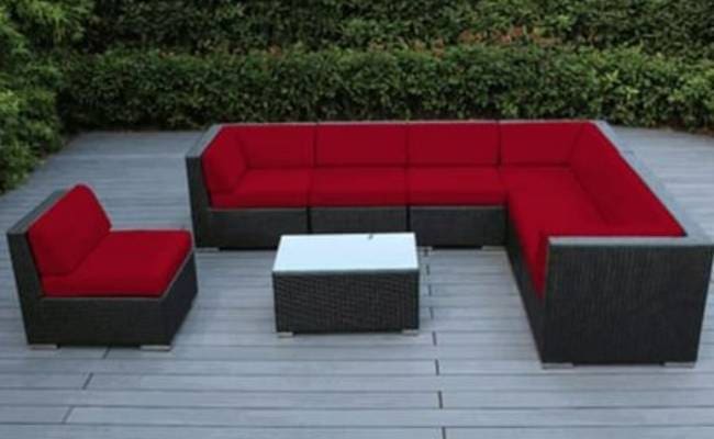 Outdoor furniture set consisting of a red sectional sofa and a single armchair with a white table, on a grey wooden deck, surrounded by green bushes.