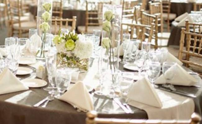 Elegant wedding reception table setting with white floral centerpieces, clear glassware, and gold chairs.