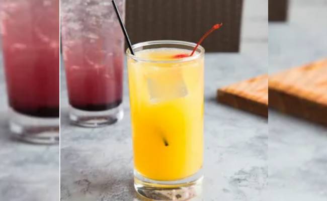 A glass of orange juice with ice and a cherry, flanked by two blurry drinks in the background, on a light gray surface.