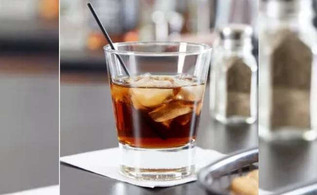 A glass of iced drink with a straw on a bar counter, accompanied by a blurred salt shaker and cutlery in the background.