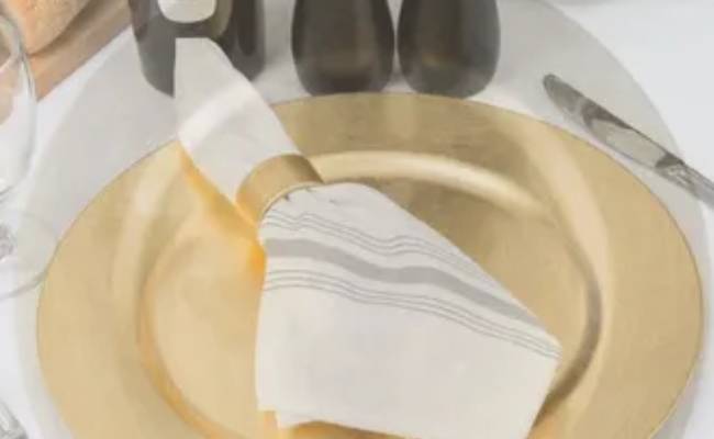 A gold charger plate with a folded white napkin and silverware, accompanied by a wine glass and black pepper grinder in the background.