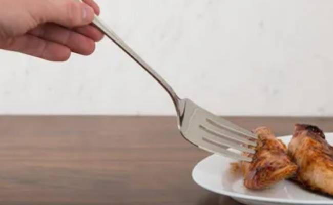 Hand using a fork to pick up a piece of grilled chicken from a white plate on a wooden table.