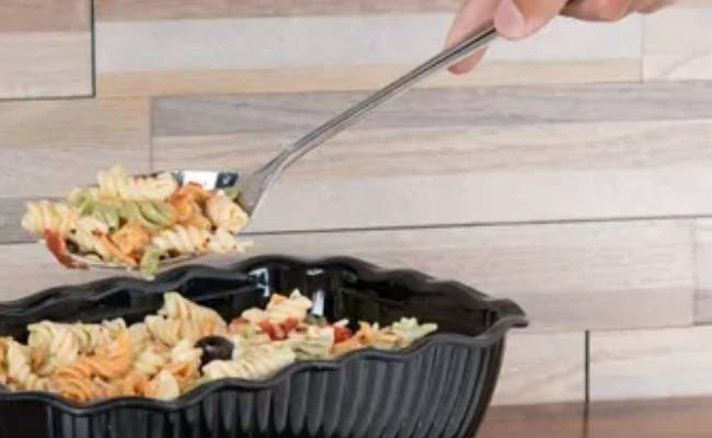 A hand using tongs to serve pasta salad from a black serving dish.