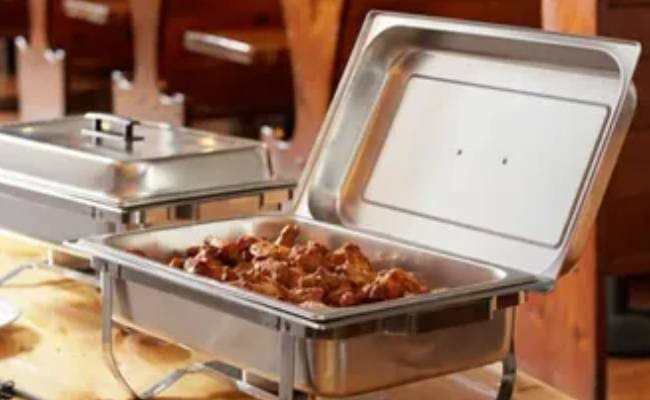 Buffet-style stainless steel chafing dishes with lids open, one containing cooked meatballs, set on a wooden table.