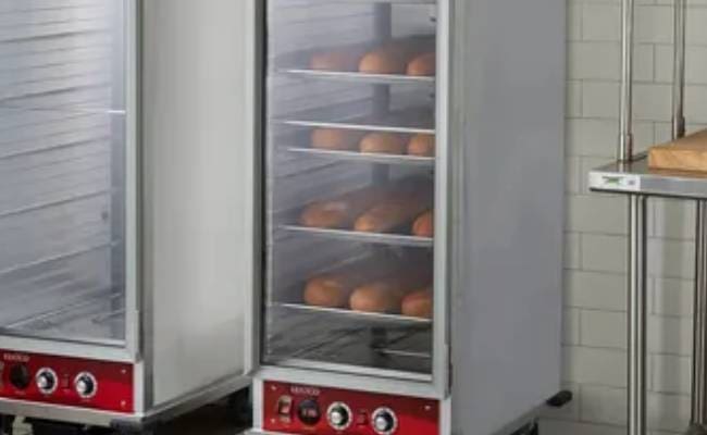 Commercial kitchen with two bread proofing cabinets, each filled with trays of bread rolls, against a tiled wall.