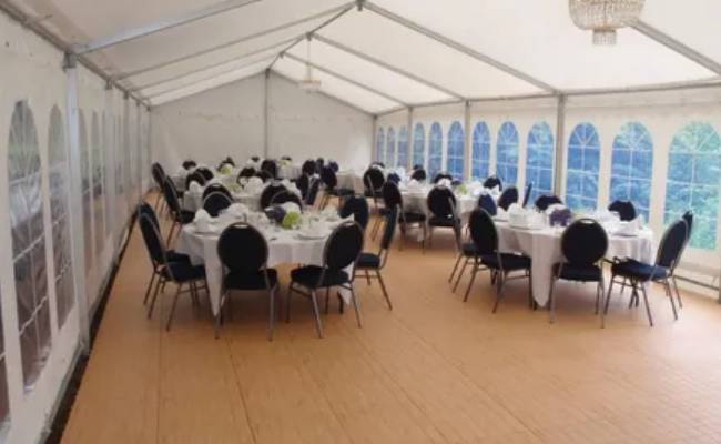 White-draped tables set for a formal event in a large tent with arched windows, unoccupied chairs facing the tables.