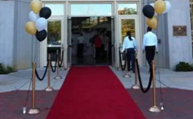 Red carpet entrance at a building with black and gold balloons tied to golden stanchions, and people walking through the open doors.