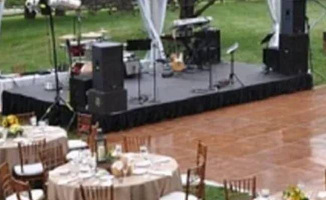 Outdoor event setup with a stage featuring musical equipment and speakers, surrounded by tables set for dining on a lawn.