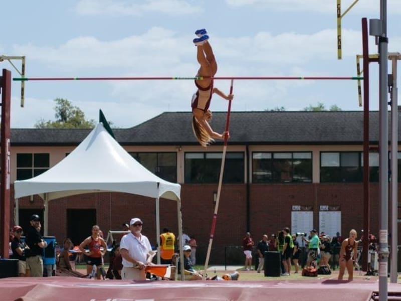 Athlete performing pole vault at an outdoor track and field event with spectators and event tents in the background.