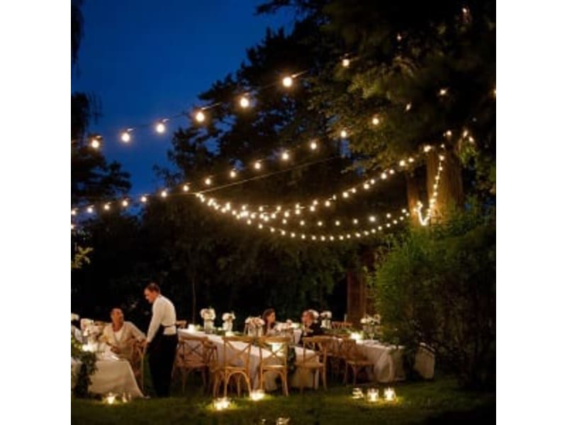 Outdoor event at dusk with string lights and elegantly decorated tables, featuring guests seated and a server attending to them.