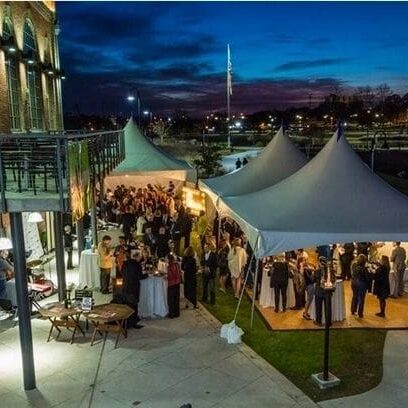 Outdoor evening event at a modern venue with people gathered under tents, adjacent to a brick building, with city lights and a dusk sky in the background.