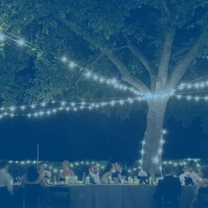 People dining outdoors at a long table under a large tree adorned with string lights at night.